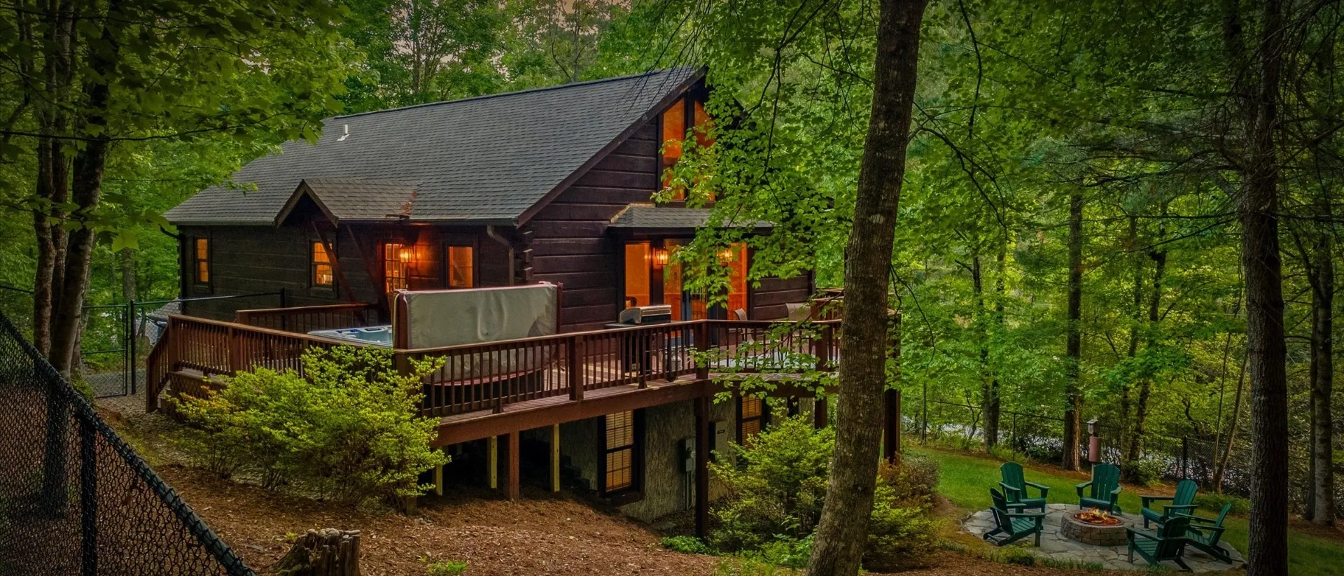 Aerial dusk view of the cabin — wraparound deck with hot tub, stone fire pit and Adirondack chairs, fully fenced wooded acre in Pisgah Forest