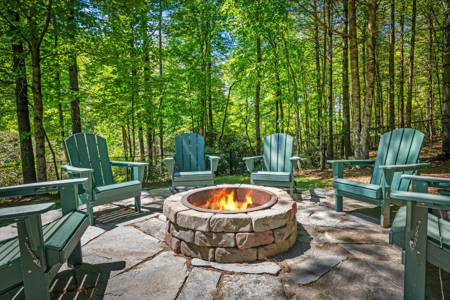 Stone fire pit ringed by sage-green Adirondack chairs on a flagstone patio, sunlit hardwood forest in the background