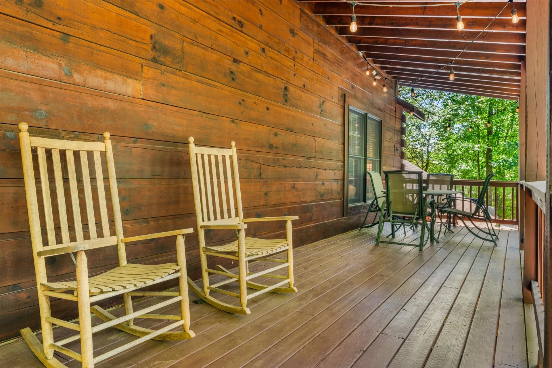 Covered front porch with two wooden rocking chairs along the cabin's stained log wall, warm string lights overhead, and forest views off the deck