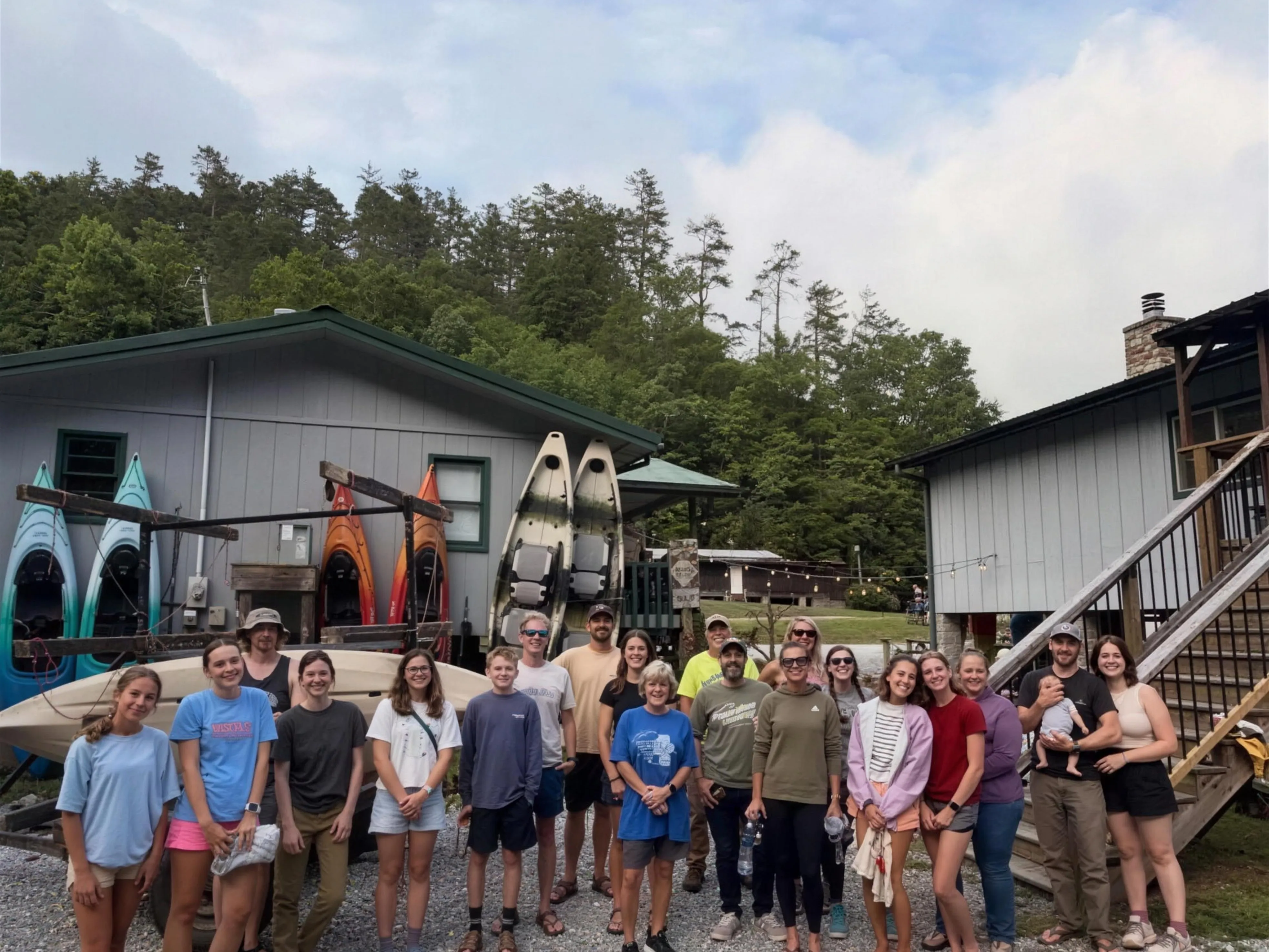 The Whitmire family and guests gathered in front of the Headwaters Outfitters shop with a fleet of canoes and kayaks racked behind them and the Blue Ridge Mountains in the background