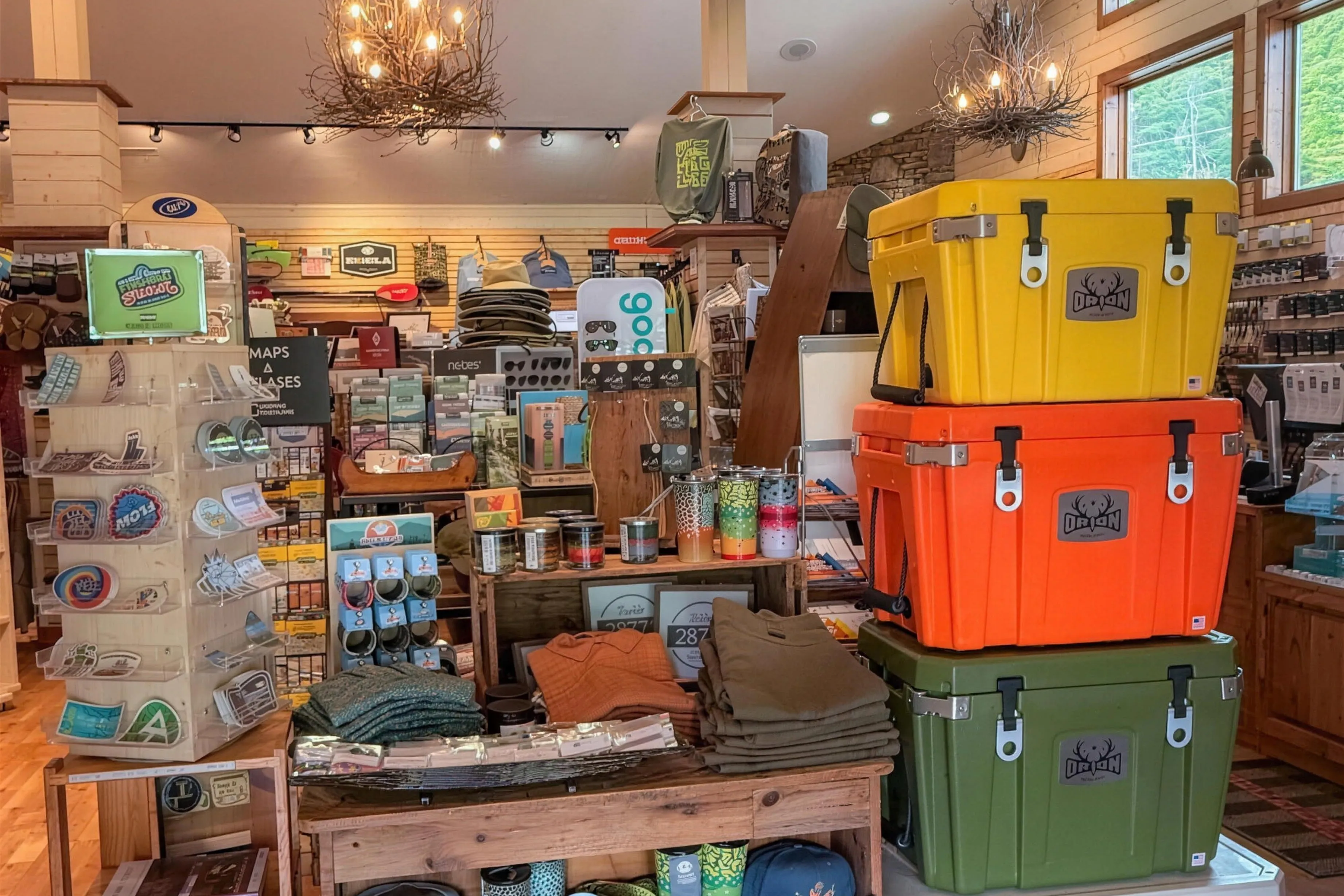 Inside the Headwaters Outfitters fly shop in Rosman — a mountain-cabin-style room with rustic chandeliers, a stone fireplace, and displays of rods, reels, hats, maps, and colorful Orion coolers