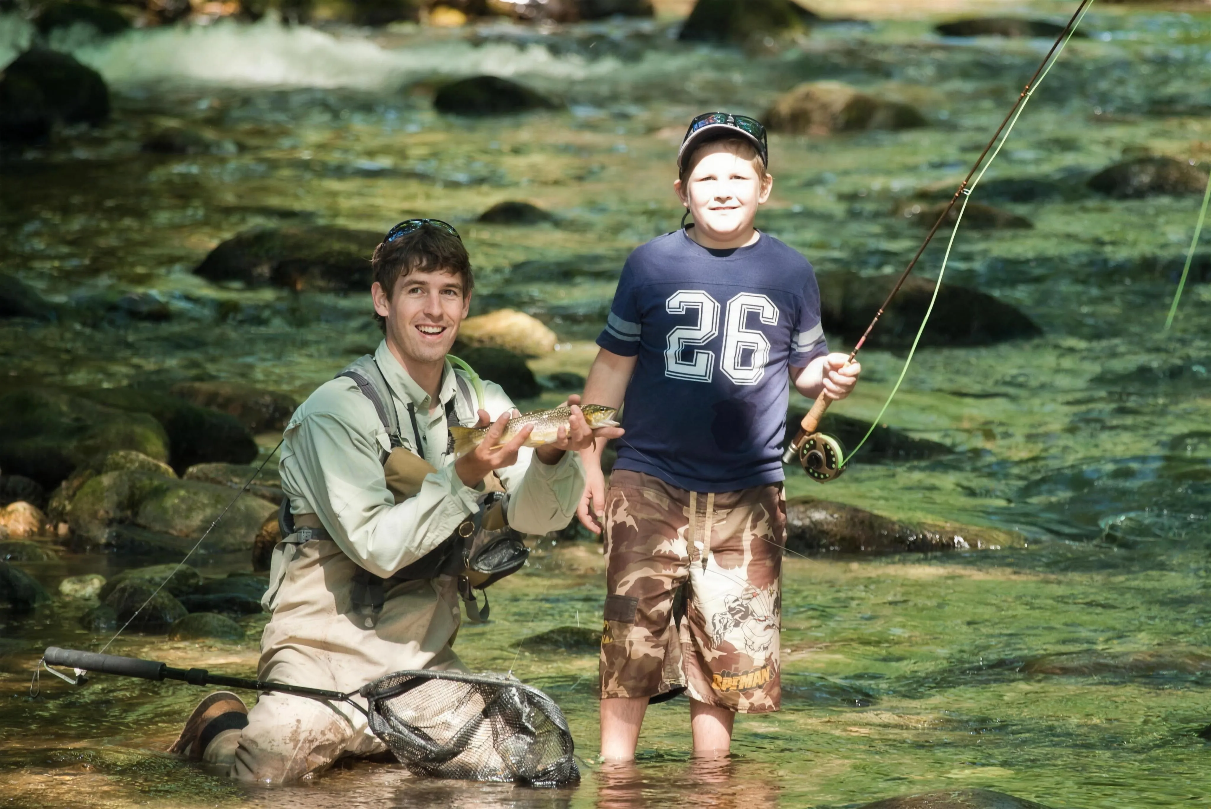 A Headwaters guide and a young angler kneeling in a clear mountain stream, the kid holding a trout caught on the fly rod he is also holding