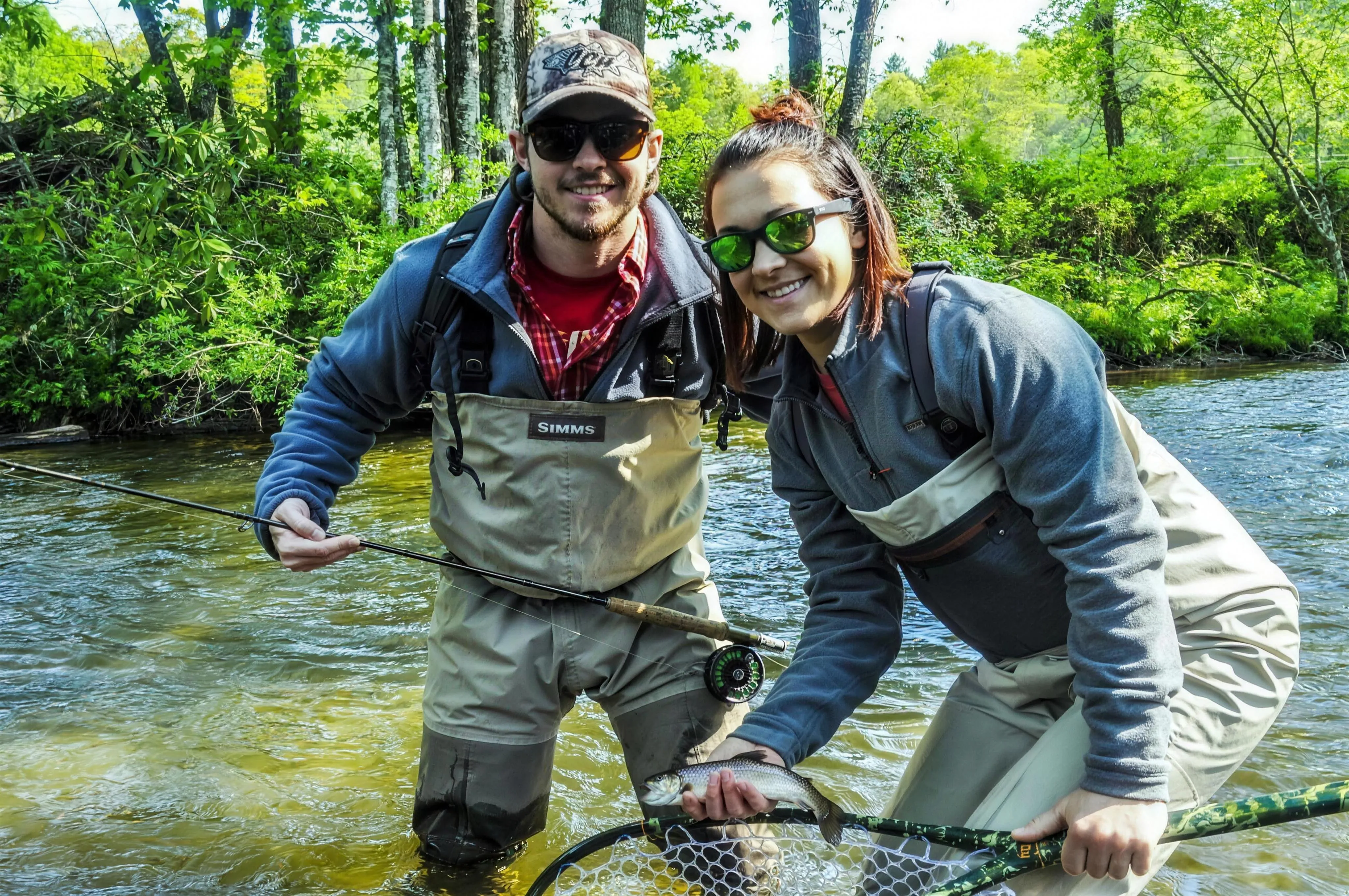 A guide and angler smile together in waders mid-stream on a trout river near Brevard, holding a fly rod ready to cast