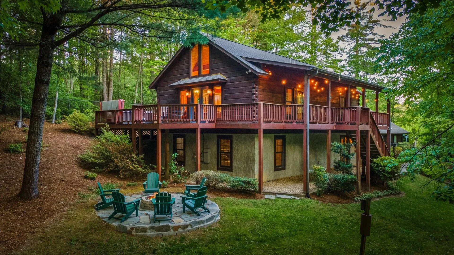 Pisgah Base Camp cabin exterior at twilight — wraparound deck, glowing string lights, and stone fire pit in the Pisgah Forest