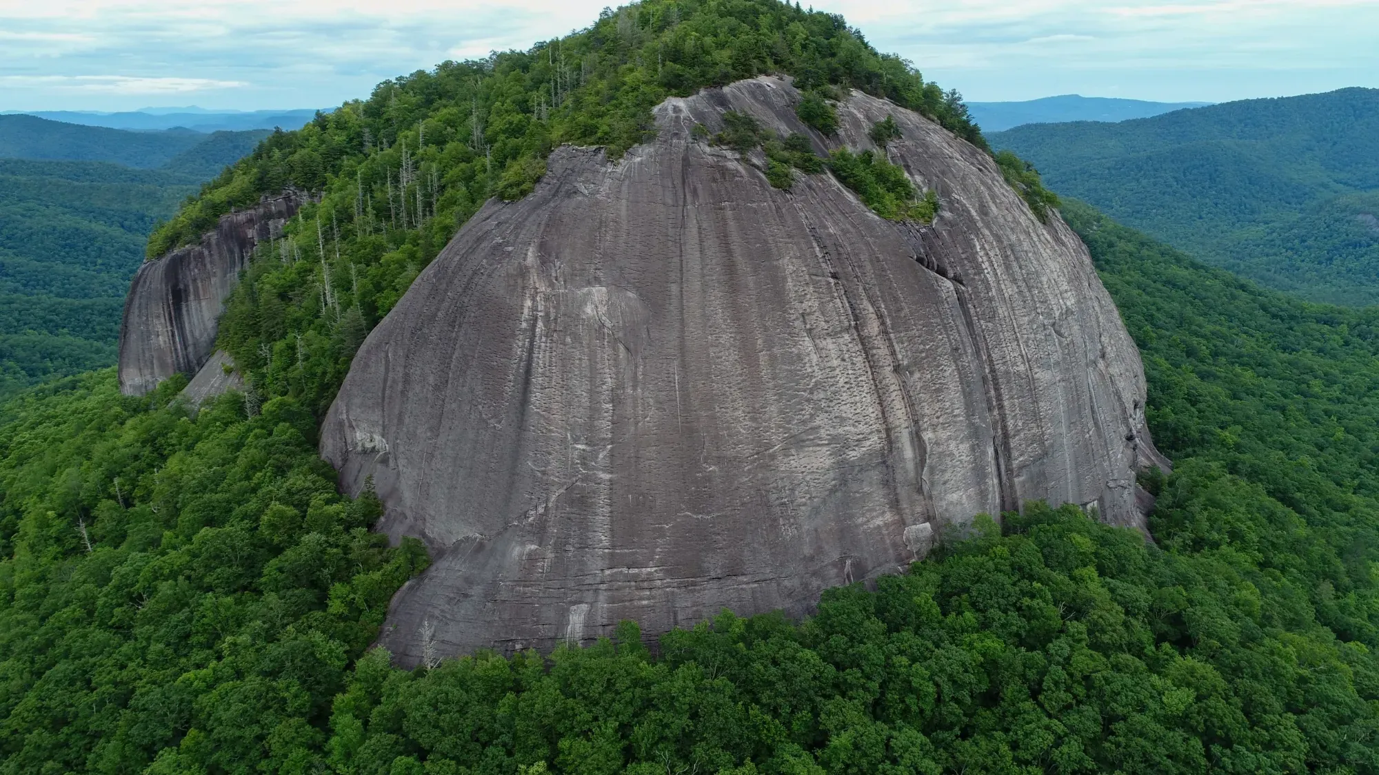 Aerial view of Looking Glass Rock, the massive granite dome the falls are named for, rising out of the green Pisgah forest with Blue Ridge mountains layered in the distance