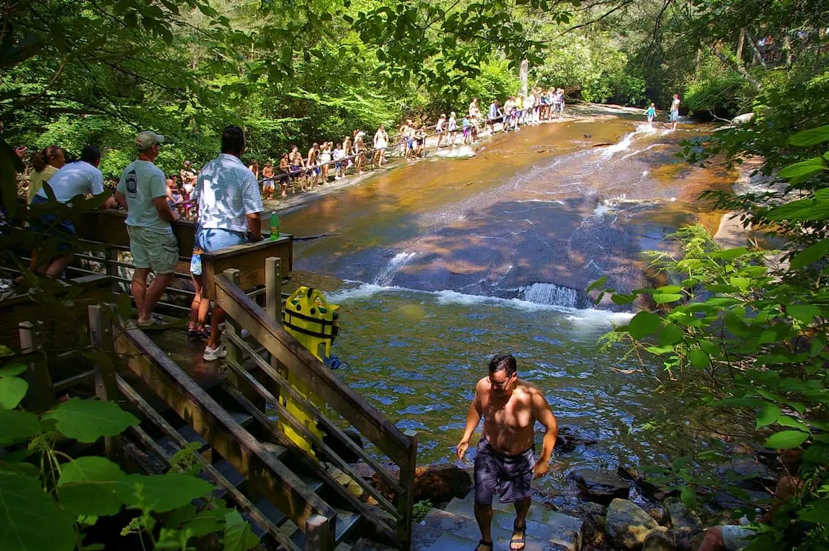 The pool and observation platform at the base of Sliding Rock, with a slider walking out of the pool while a crowd lines up at the top of the granite chute